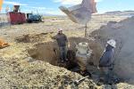 Two men stand nearly shoulder deep in a large hole in the ground watching a heavy load dangles on a chain from a loader bucket. Large pile of dirt and other equipment in the background.