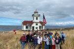 A group of volunteers are raising the American flag. They are standing in tall yellow grass with a white lighthouse in the background. 