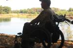 A photo showing a man in a wheelchair next to a river. He’s wearing camouflage and has an adaptive bow mount on his wheelchair.