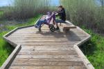 A photo showing a wooden boardwalk leading away from the viewer. A teenage boy sits on a wooden bench on the widened part of the boardwalk with his younger sister who is sitting in a mobility device.