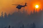 A helicopter flies over a smoked filled forest in an orange sky.